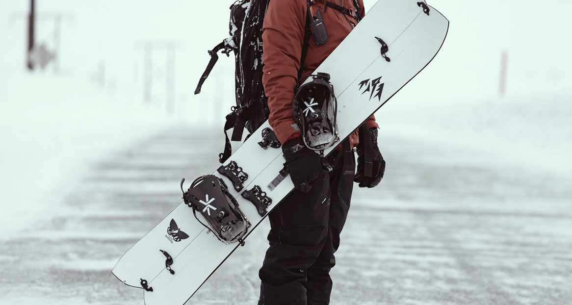 Eine Person in Winterkleidung hält ein Splitboard mit Bindungen in einer verschneiten Umgebung.