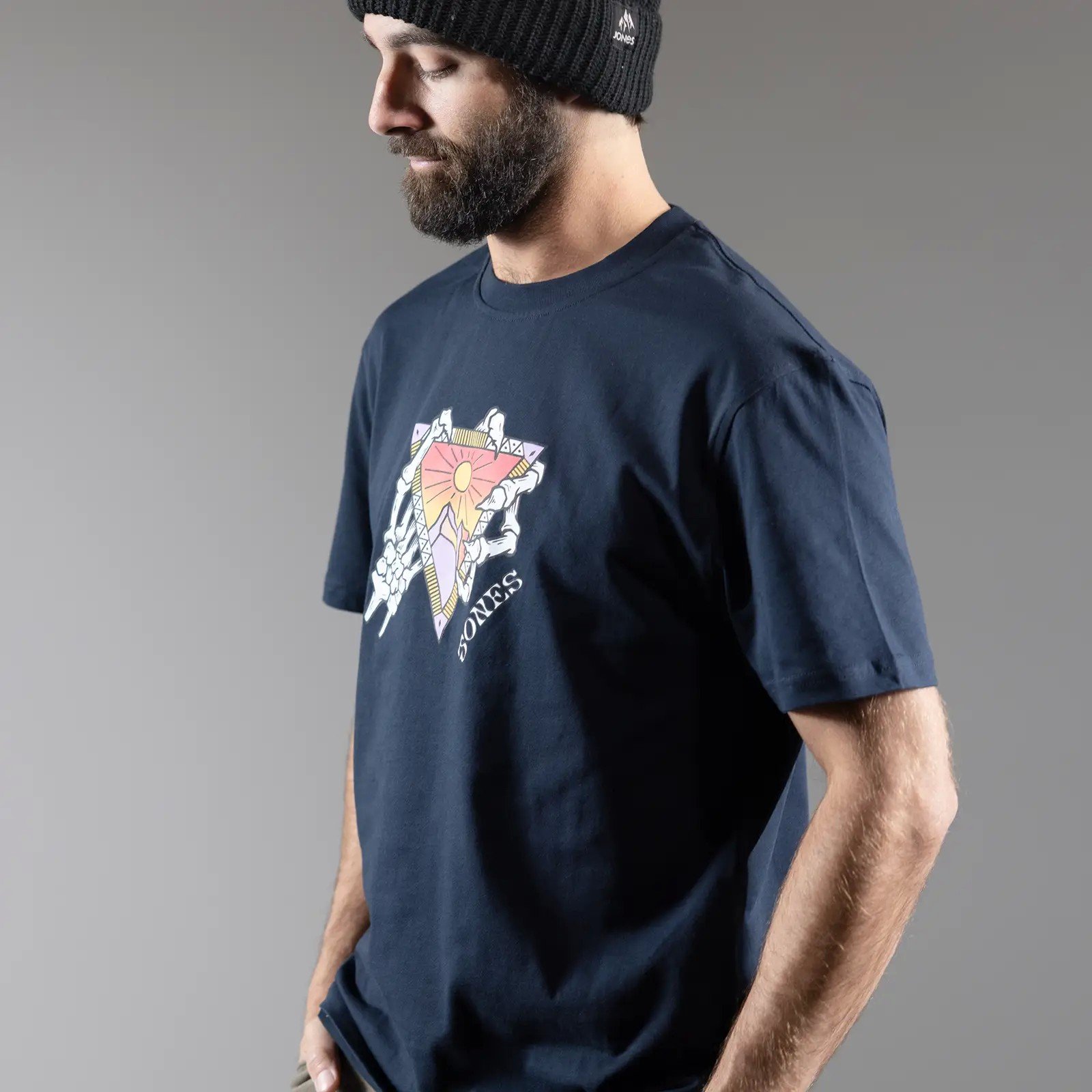 Mann im Jones T-Shirt Mann mit Mütze trägt Jones Organic Cotton Tee mit Grafik eines Dreiecks und Skeletthänden.