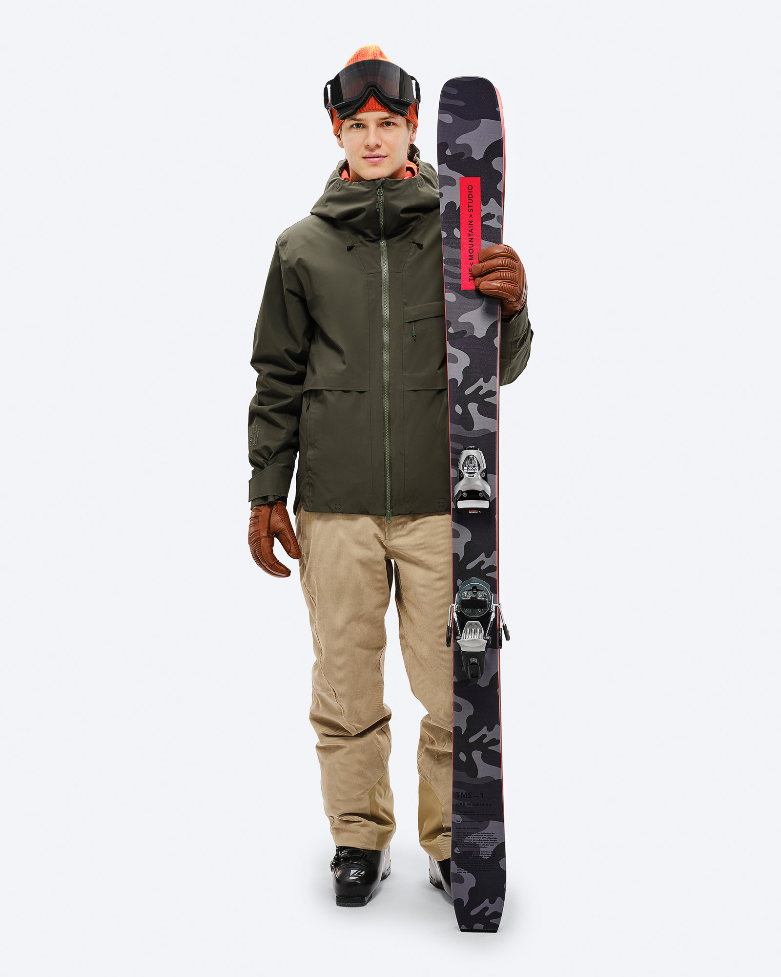 Skijacke S-3 Carv Grün Model mit S-3 Carv Insulated Stretch Jacket in Grün hält Ski mit Tarnmuster, trägt Mütze und Skibrille.