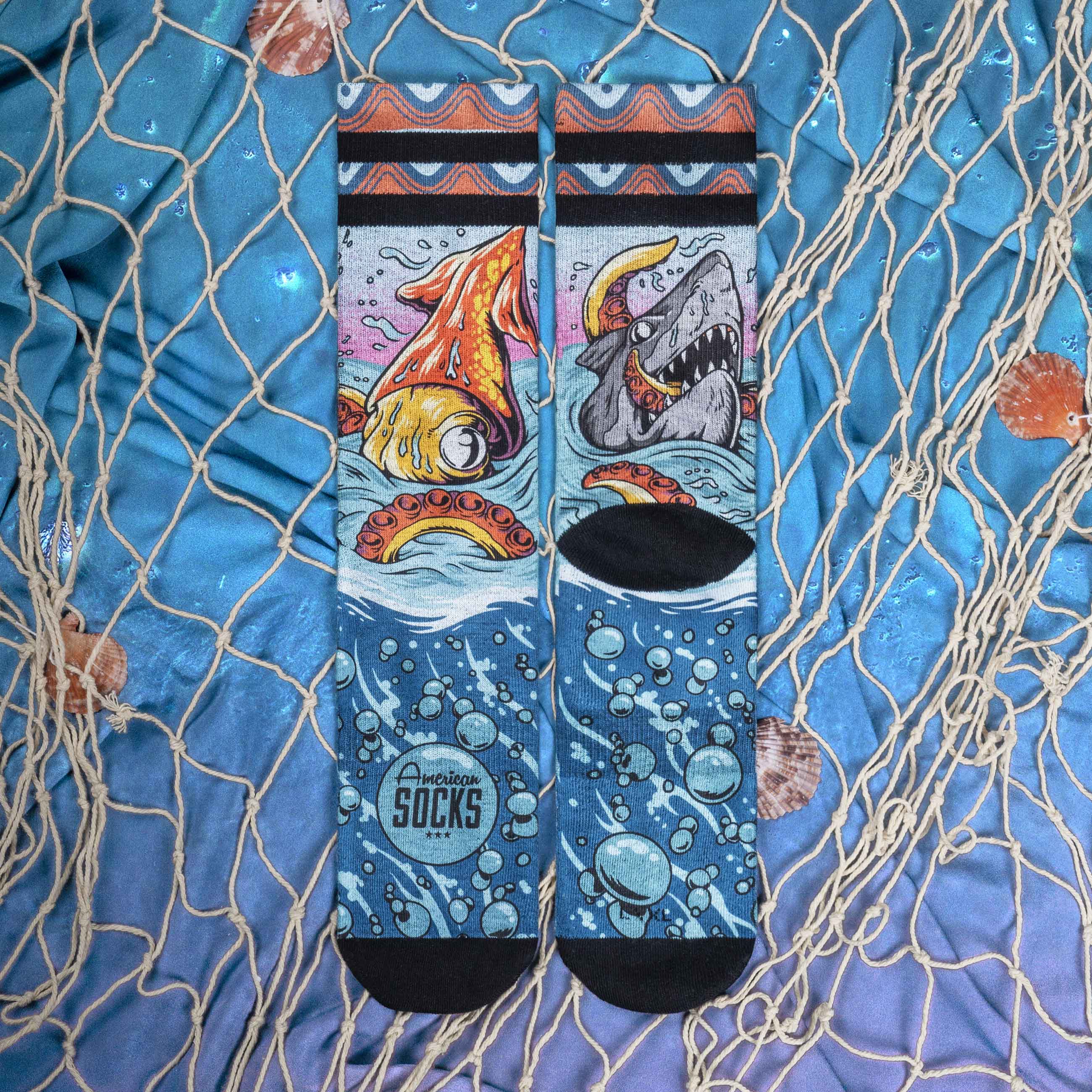 Chaussettes Seamonster bleues Deux chaussettes 'Seamonsters Mid High' avec pieuvre, requin, bulles et logo 'American Socks' sur tissu bleu avec coquillages et filet de pêche.
