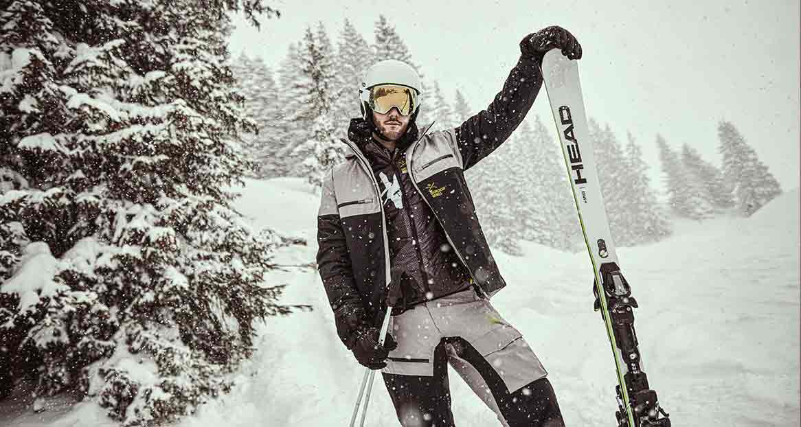 Ein Skifahrer in Winterkleidung steht vor verschneiten Bäumen und hält einen Ski.