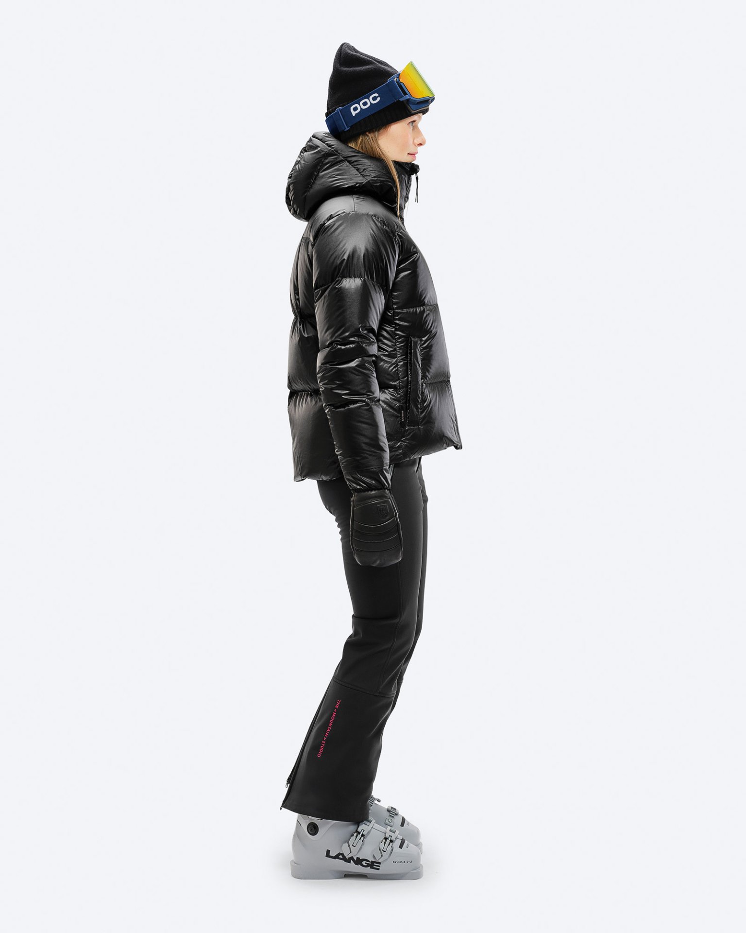 Skifahrerin in Schwarz Frau in schwarzer Skihose und Jacke von der Seite, mit Skischuhen, Mütze und Skibrille.