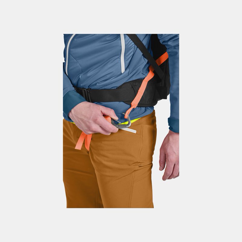 Beingurt am Rucksack Detailaufnahme: Person befestigt Beingurt am Ortovox Avabag Litric Tour 30 Rucksack. Der Gurt ist orange, die Hose braun.