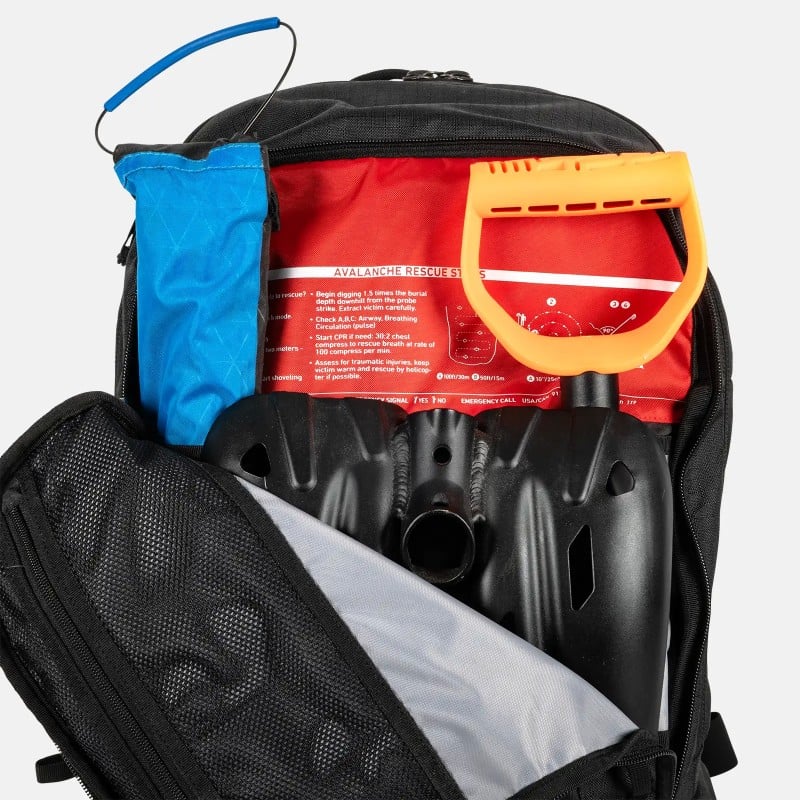 Offener Jones Rucksack 25L Offener Jones Further 25L Rucksack mit Lawinenschaufel, Sonde und Erste-Hilfe-Informationen in Rot. Fach mit Netzstruktur sichtbar.