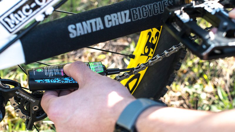 Fahrrad-Kette wird mit Muc-Off Wet Lube geschmiert. Flasche mit grünem Etikett. Fahrrad-Details sichtbar.