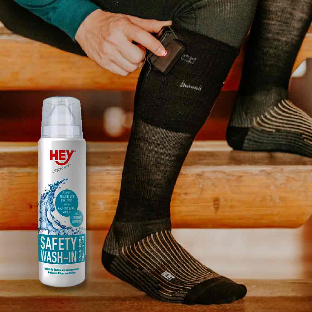 Wie wasche ich beheizbare Socken? Eine Person befestigt ein Akku an einer schwarzen Heizsocke, neben Waschmittel "HEY Sport Safety Wash-In".