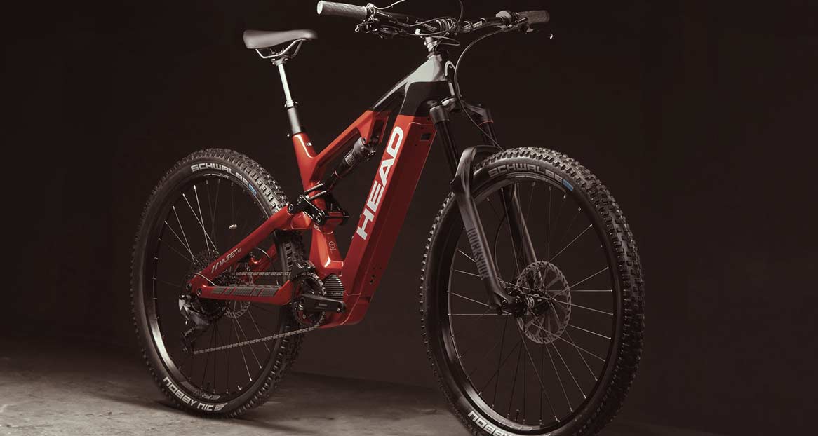 Ein rotes und schwarzes E-Mountainbike der Marke Head steht vor einem dunklen Hintergrund, das Licht reflektiert.