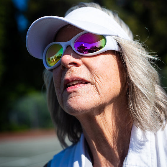 Femme âgée aux cheveux gris et à la visière blanche, portant des lunettes de soleil colorées The Slammer Miami Nights.