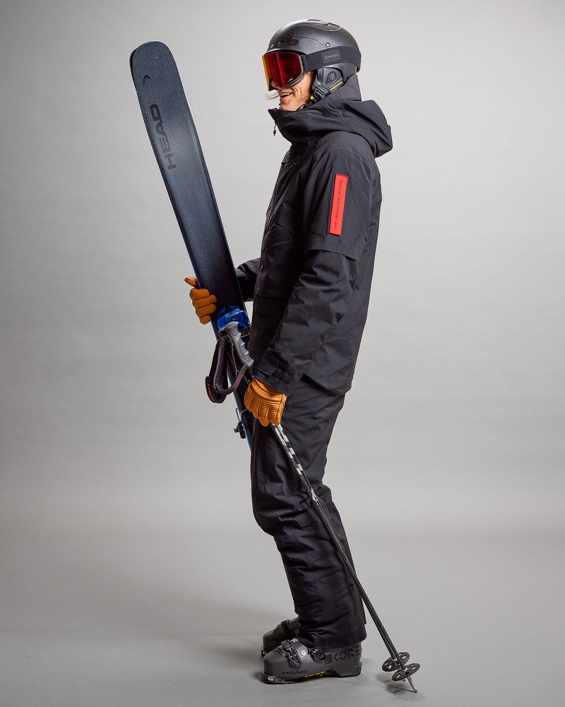 Skifahrer in Gore Tex Person in schwarzer Gore Tex Skijacke, Hose, Helm und Skibrille hält Ski und Skistöcke.