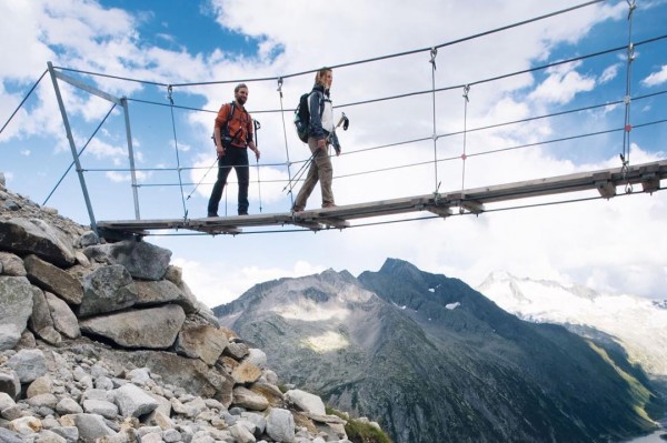 Hochwertigen Bergschuhe kaufen | ski-shop.ch Zwei Wanderer auf einer Hängebrücke vor Bergpanorama unter blauem Himmel mit Wolken. Steine im Vordergrund.