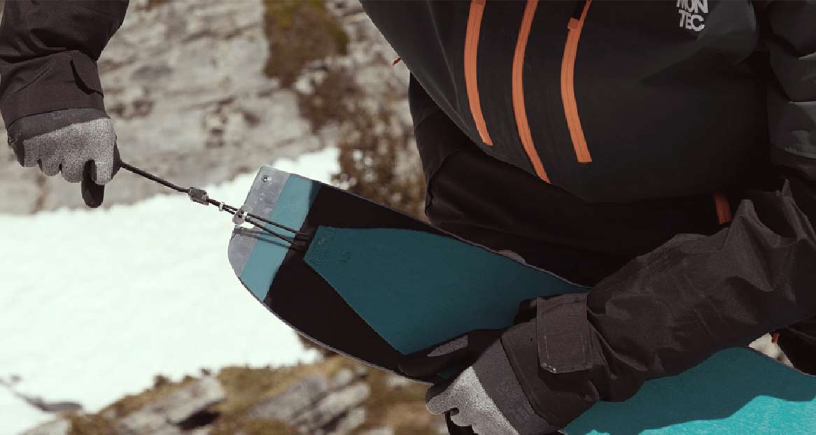 Eine Person hantiert mit Splitboard-Fellen. Die Felle sind türkis und schwarz und mit einer Schnur verbunden. Schnee im Hintergrund.
