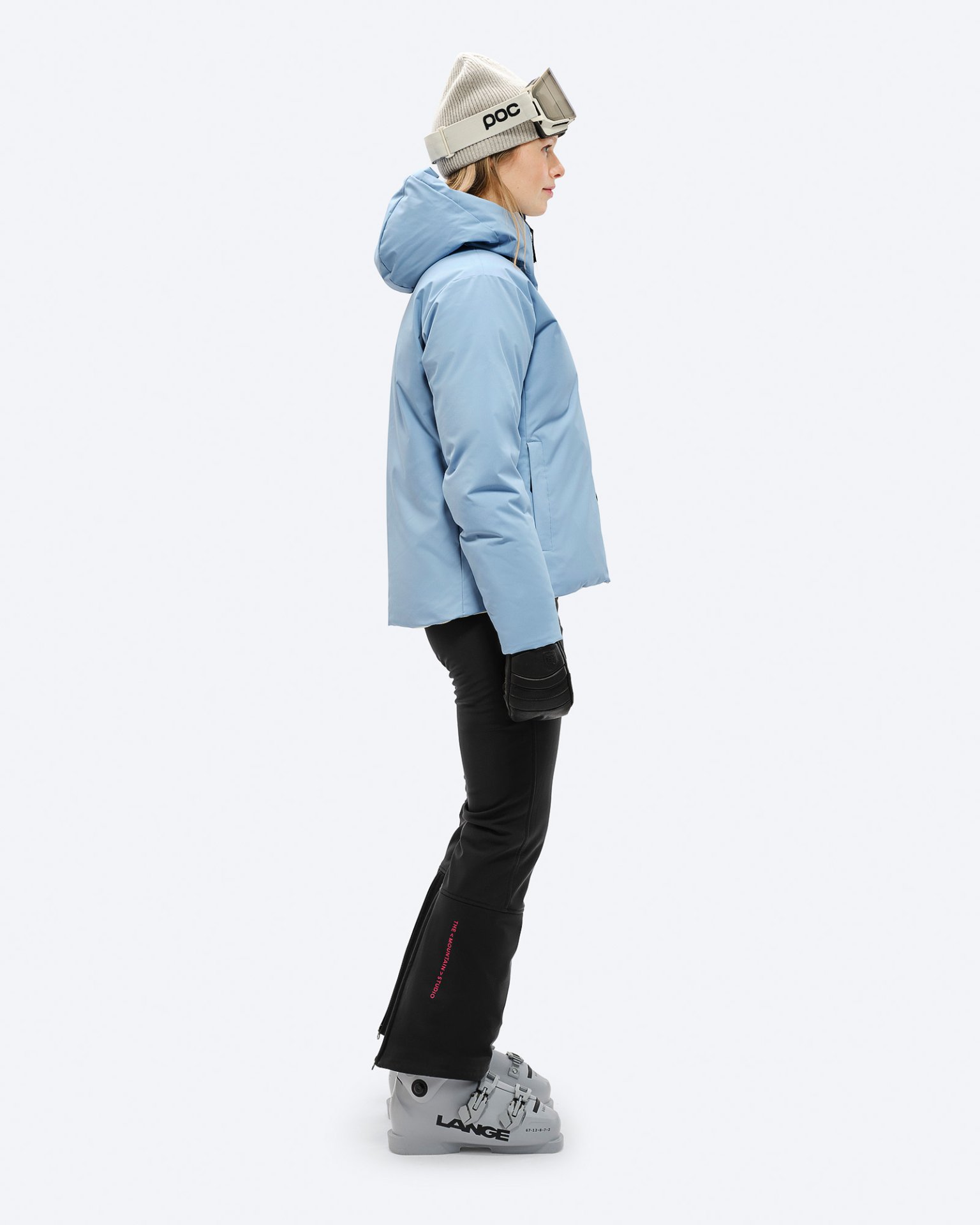 Ski-Bekleidung für Frauen Frau in Ski-Bekleidung, inklusive hellblauer Daunenjacke, Skibrille und Mütze, seitliche Ansicht vor neutralem Hintergrund.