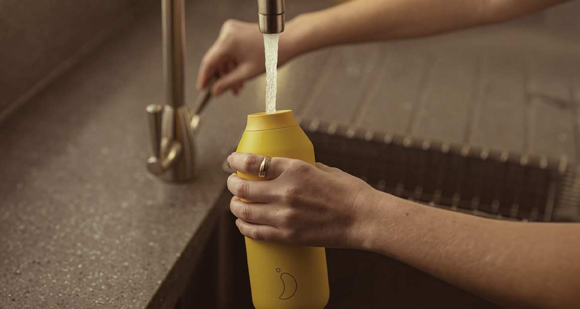 Eine gelbe Trinkflasche wird unter einem Wasserhahn in einer Spüle mit Wasser befüllt. Die Flasche hat ein tropfenförmiges Logo.