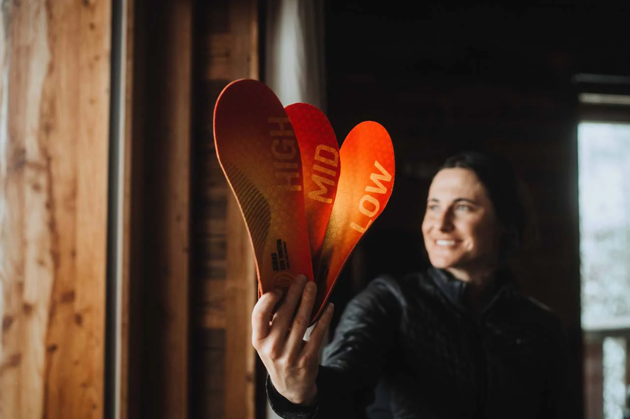 3Feet® Eco Skisohlen: Umweltfreundlicher Komfort für Skifahrer Frau zeigt drei rot-gelbe Einlegesohlen mit Aufschriften "HIGH", "MID" und "LOW" in die Kamera.
