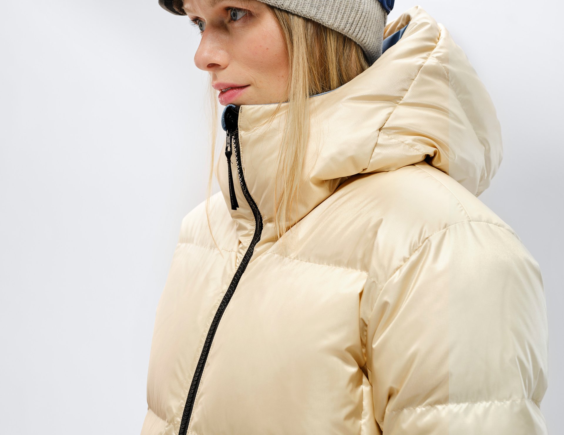 Damen Daunenjacke mit Mütze Profil einer Frau mit beiger Daunenjacke und Mütze. Die Jacke hat einen schwarzen Reißverschluss und eine Kapuze.