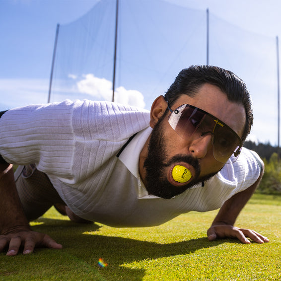 Homme en position de pompes avec une balle de golf jaune dans la bouche et des lunettes de soleil The Exec.