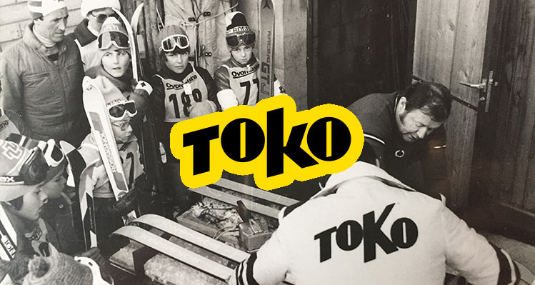 Schwarz-Weiß-Aufnahme einer Gruppe Skifahrer, einige Kinder mit Skibrillen und Rennanzügen, und zwei Erwachsene, die Skier präparieren, mit dem "TOKO"-Logo überlagert.