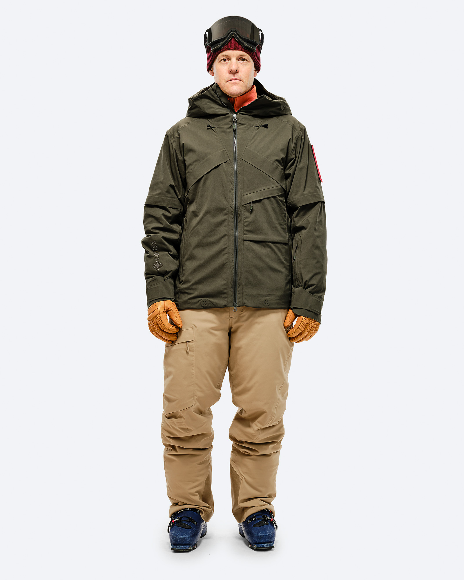 Skifahrer Outfit Person in Skikleidung: Olivgrüne Jacke, sandfarbene Hose und Skischuhe vor weißem Hintergrund.