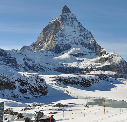 Ein schneebedeckter Berg mit einem spitz zulaufenden Gipfel, umgeben von einer Winterlandschaft mit einem kleinen See im Vordergrund.