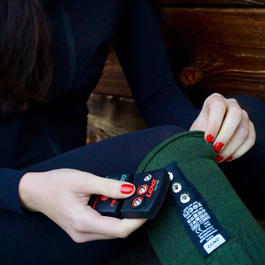 Eine Person sitzt mit schwarzen Leggings, hält schwarze Akkus mit roten Knöpfen und eine grüne Socke mit Lenz-Logo in der Hand.