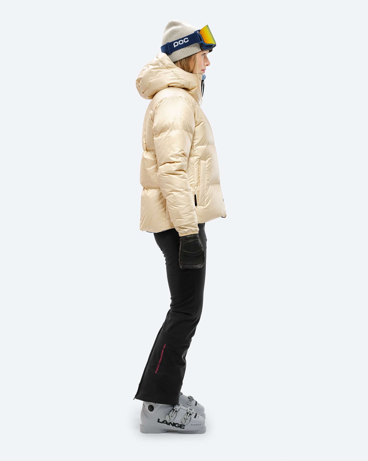 Damen Winterbekleidung Frau in beiger Daunenjacke mit Kapuze, Skihose und Skischuhen in Seitenansicht. Winterbekleidung.