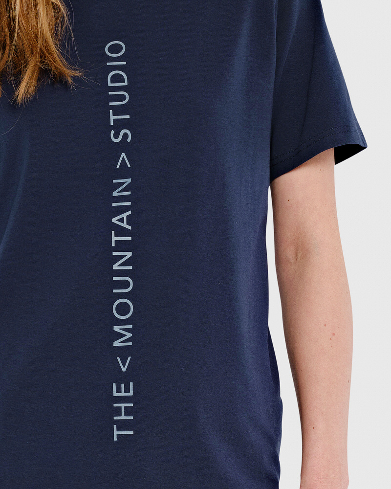 Dunkelblaues T-Shirt mit vertikalem, hellgrauem Logo-Druck: "THE < MOUNTAIN > STUDIO".