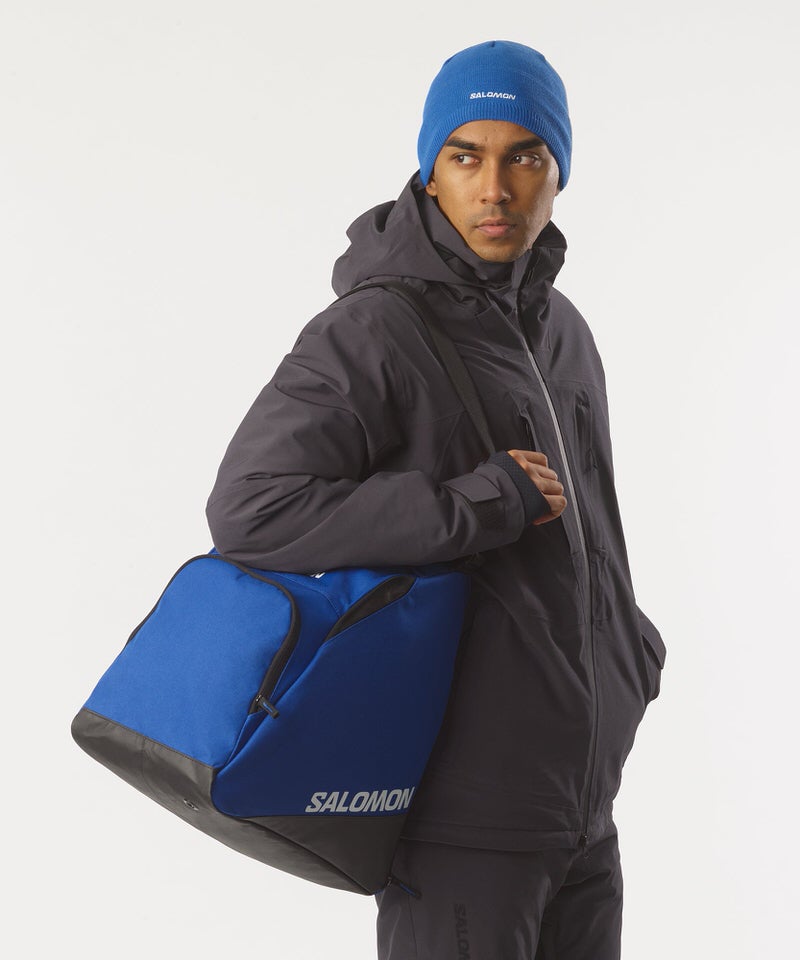 Mann mit blauer Mütze, trägt Salomon Skischuhtasche über der Schulter. Die Tasche ist blau-schwarz mit Salomon Logo.
