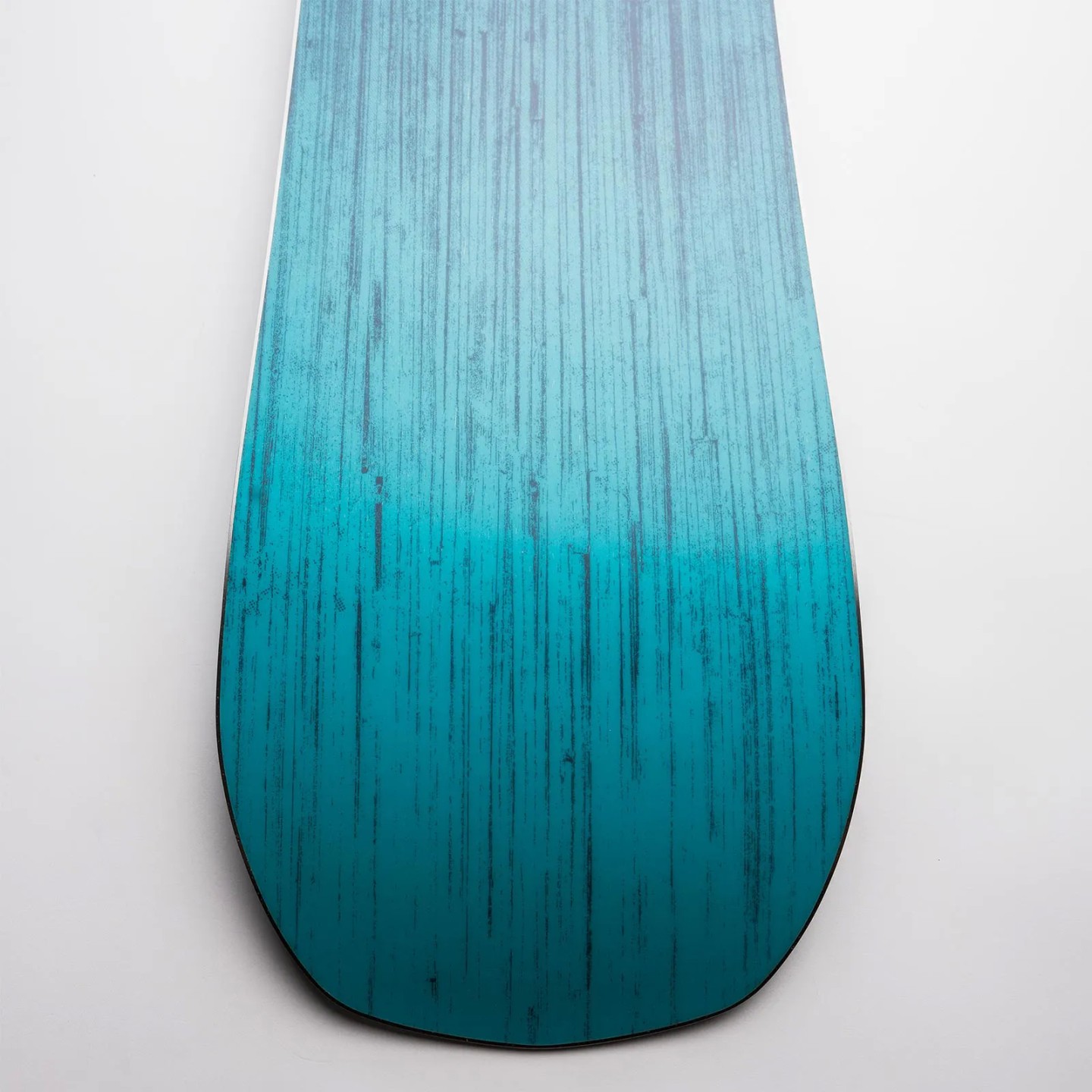 Nidecker Score Snowboard Detailansicht vom Nidecker Score Snowboard: Das Board hat ein blau-türkises Farbverlaufsmuster mit dunklen, vertikalen Linien.