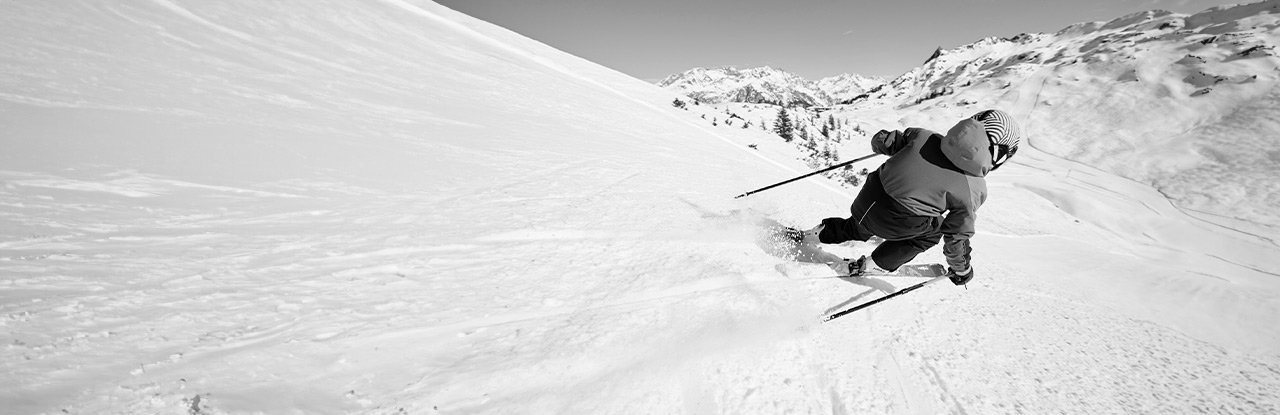 Skifahren im Schnee Schwarzweißaufnahme einer Person beim Skifahren auf einer verschneiten Piste, mit Bergen im Hintergrund.