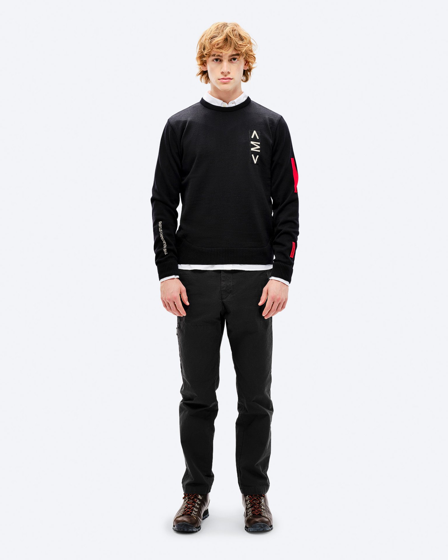 Mann im K-3 Ski Sweatshirt Mann mit rotem Haar trägt schwarzen K-3 Ski Sweater mit roten Akzenten und schwarzen Hosen, posiert vor einem weißen Hintergrund.