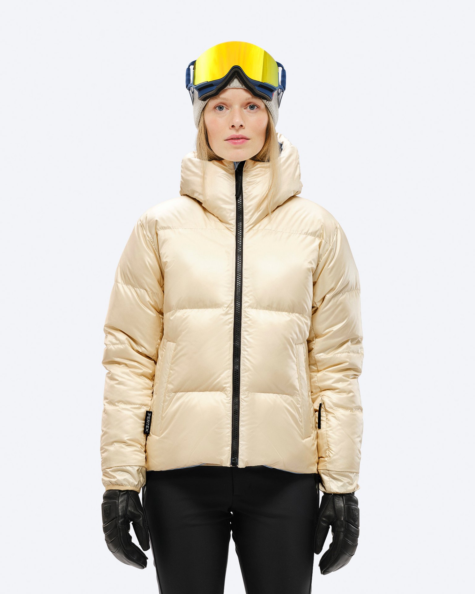 Beige Daunenjacke mit Kapuze Eine Frau trägt eine glänzende, beige Daunenjacke mit Kapuze, schwarze Handschuhe und Skibrille vor einem weißen Hintergrund.