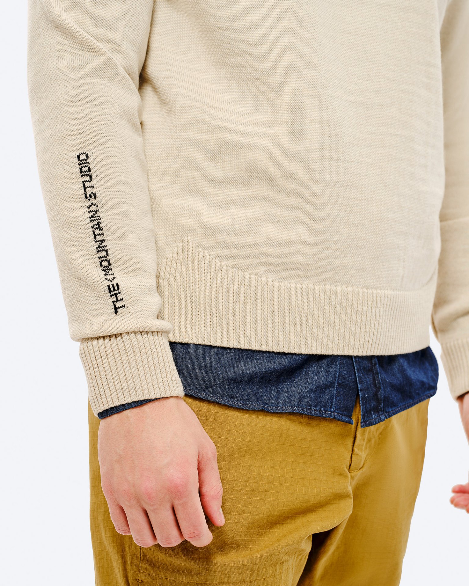 St. Moritz Ski Sweater Detailansicht vom St. Moritz Ski Sweater, beige, mit Schriftzug auf dem Ärmel, über einem blauen Hemd und beigen Hosen.