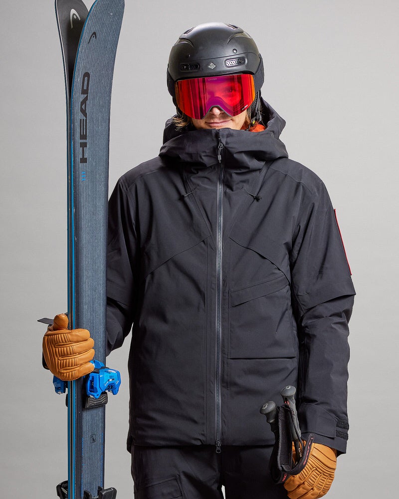 Skifahrer mit Ausrüstung Person in schwarzer Gore-Tex-Skijacke mit Ski, Helm und Brille, hält Stöcke und trägt Handschuhe.