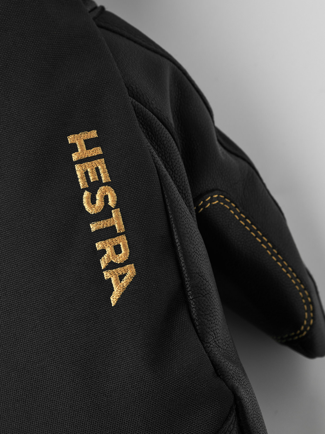 Schwarze Hestra-Handschuhe Detailansicht eines schwarzen Hestra-Handschuhs mit goldener 'HESTRA'-Stickerei und gelben Nähten.