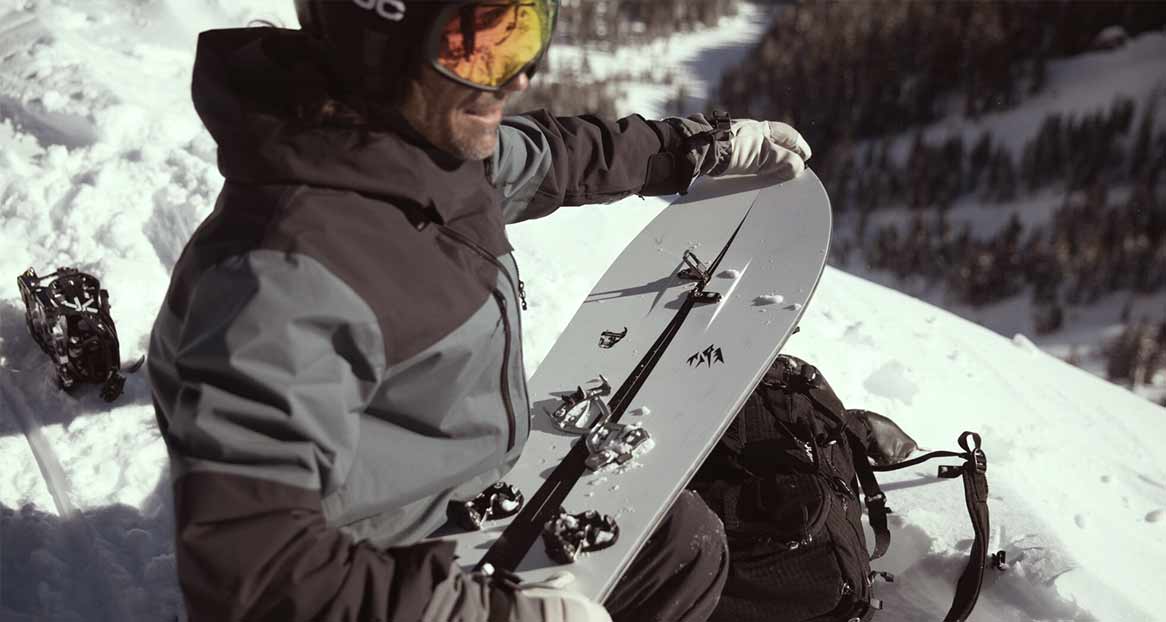 Mann mit Splitboard in verschneiter Berglandschaft. Er trägt Helm, Skibrille und Winterkleidung. Rucksack und Bindung im Schnee.