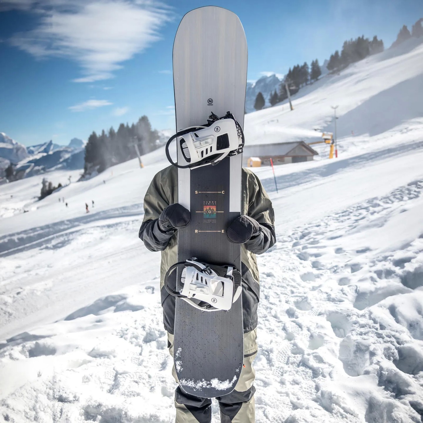 Nidecker Score Snowboard Person hält ein Nidecker Score Snowboard mit montierten Bindungen vor dem Körper; verschneite Bergkulisse.
