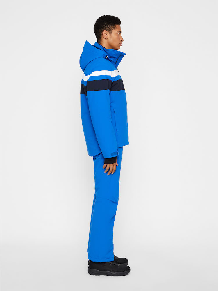 J.Lindeberg Skijacke Herren Ein Mann trägt eine blaue J.Lindeberg Franklin 2L Skijacke und Hose im Profil. Die Jacke hat weiße und marineblaue Streifen.
