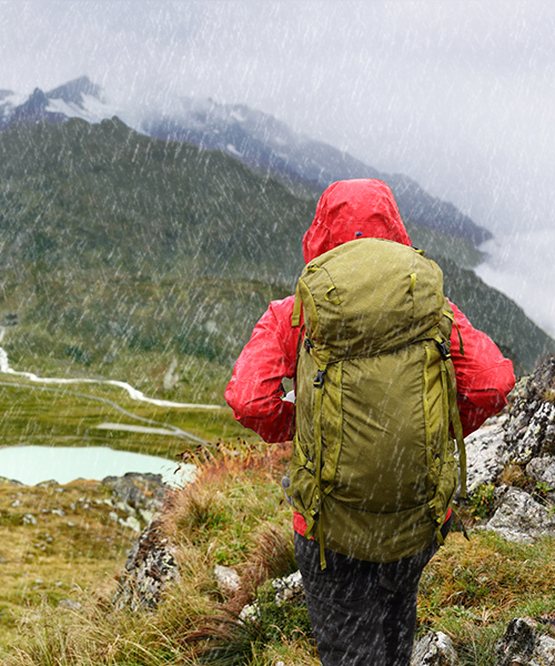 HEY SPORT® MICRO WASH Wanderer in roter Jacke mit Rucksack im Regen vor Berglandschaft mit See.