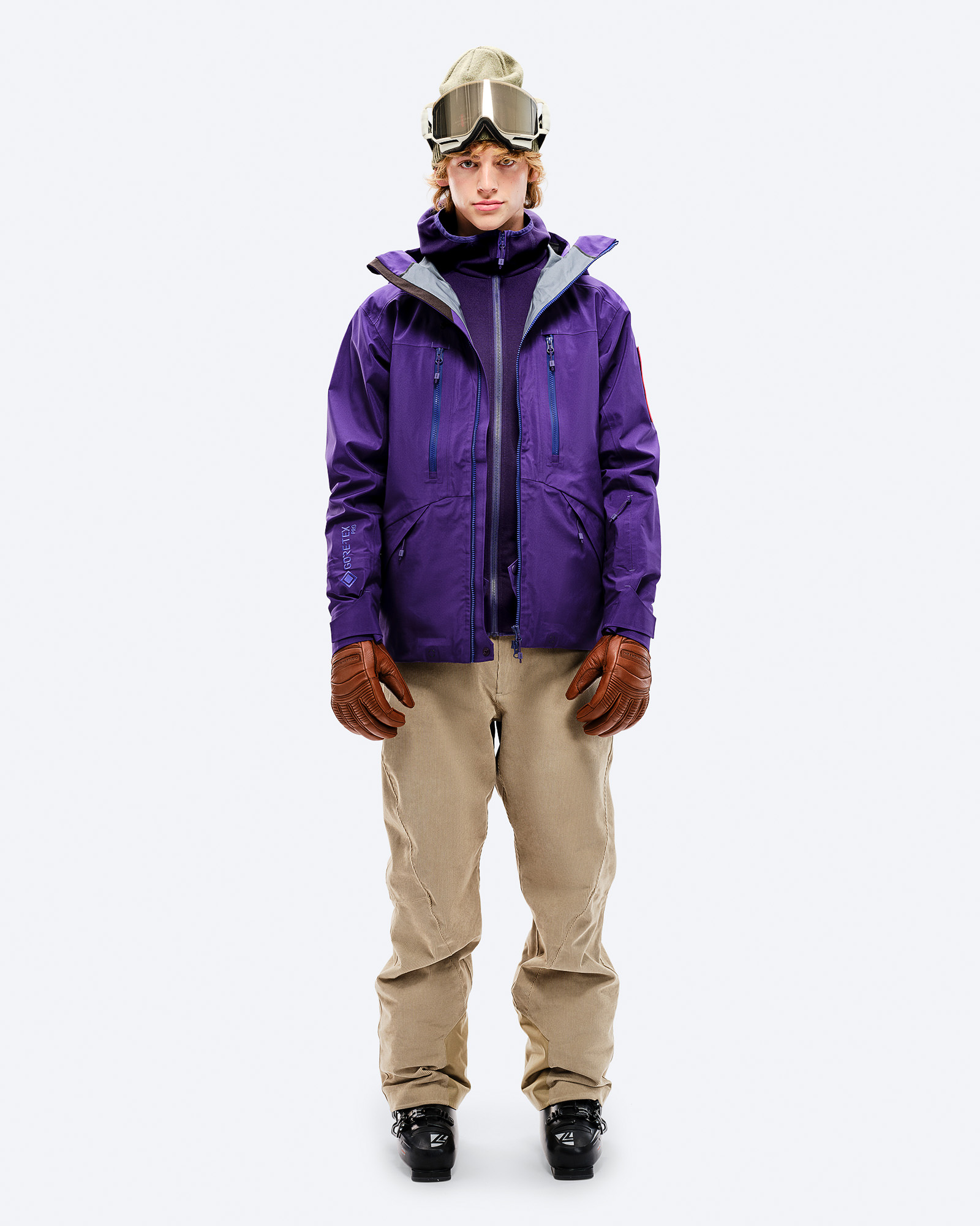 Violette GORE-TEX Jacke Mann in violetter GORE-TEX EPE 3L Shell Jacket, beigen Hosen, Skischuhen und Brille, frontal auf Weiß.