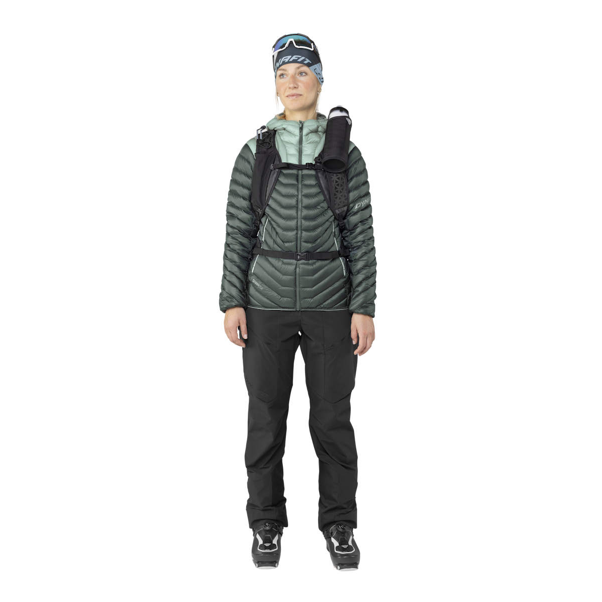 Eine Frau in Ski-Bekleidung, mit Dynafit Ridge Ultralight Daunenjacke in Grün, Hose und Skischuhen.