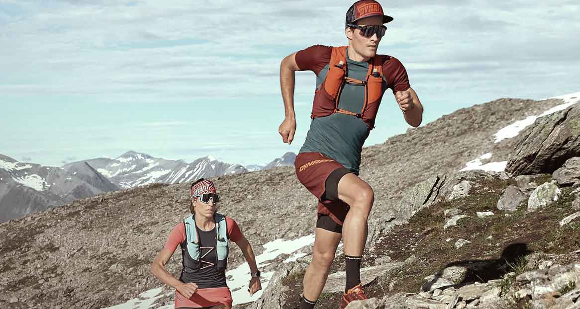 Zwei Trailrunner in Sportbekleidung laufen bergauf. Im Hintergrund sind schneebedeckte Berge und ein bewölkter Himmel sichtbar.