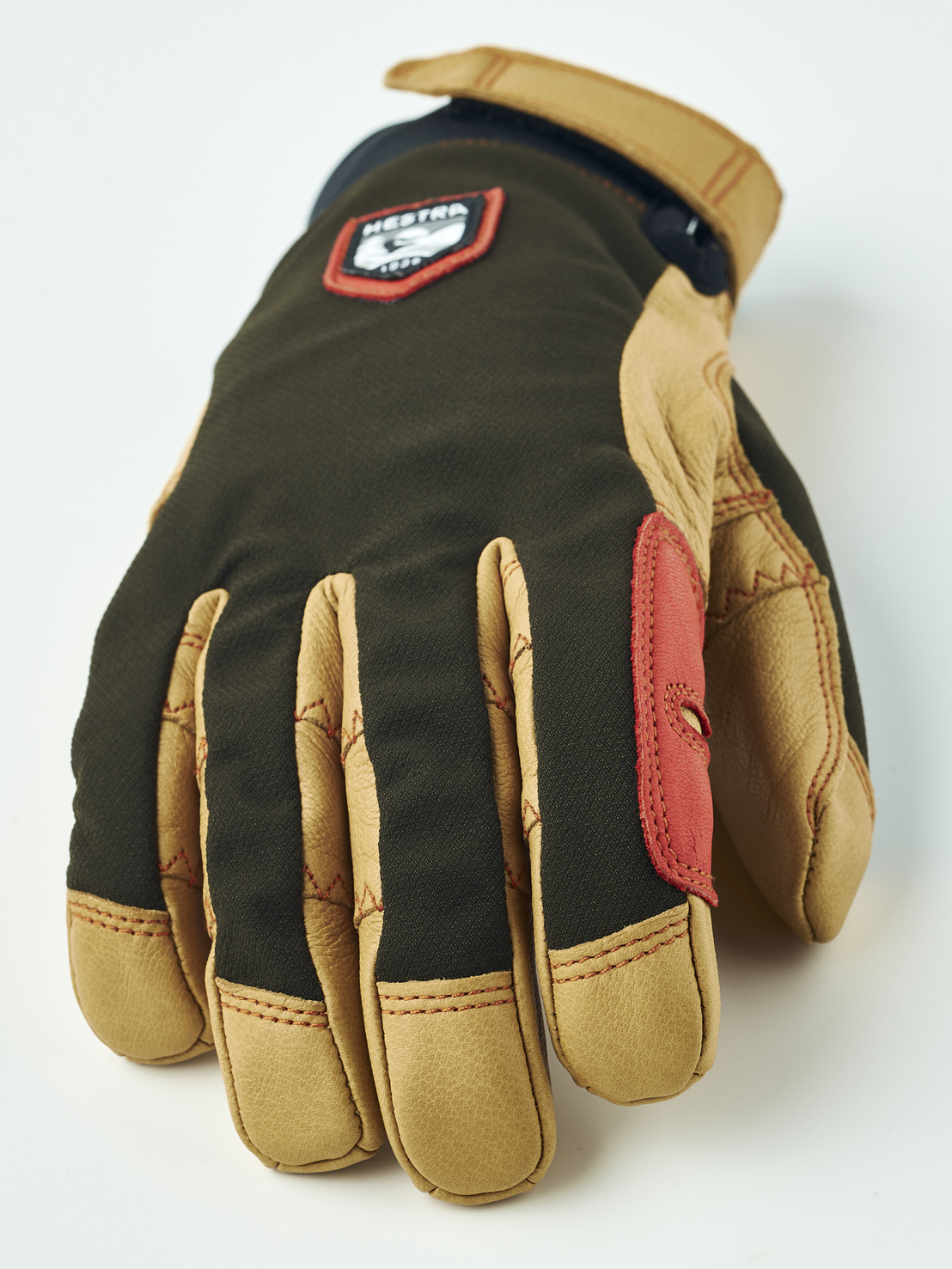 Hestra Handschuh Braun Nahaufnahme eines braunen Hestra-Handschuhs mit beigen Lederdetails und rotem Patch. Das Markenlogo ist auf dem Handrücken zu sehen.