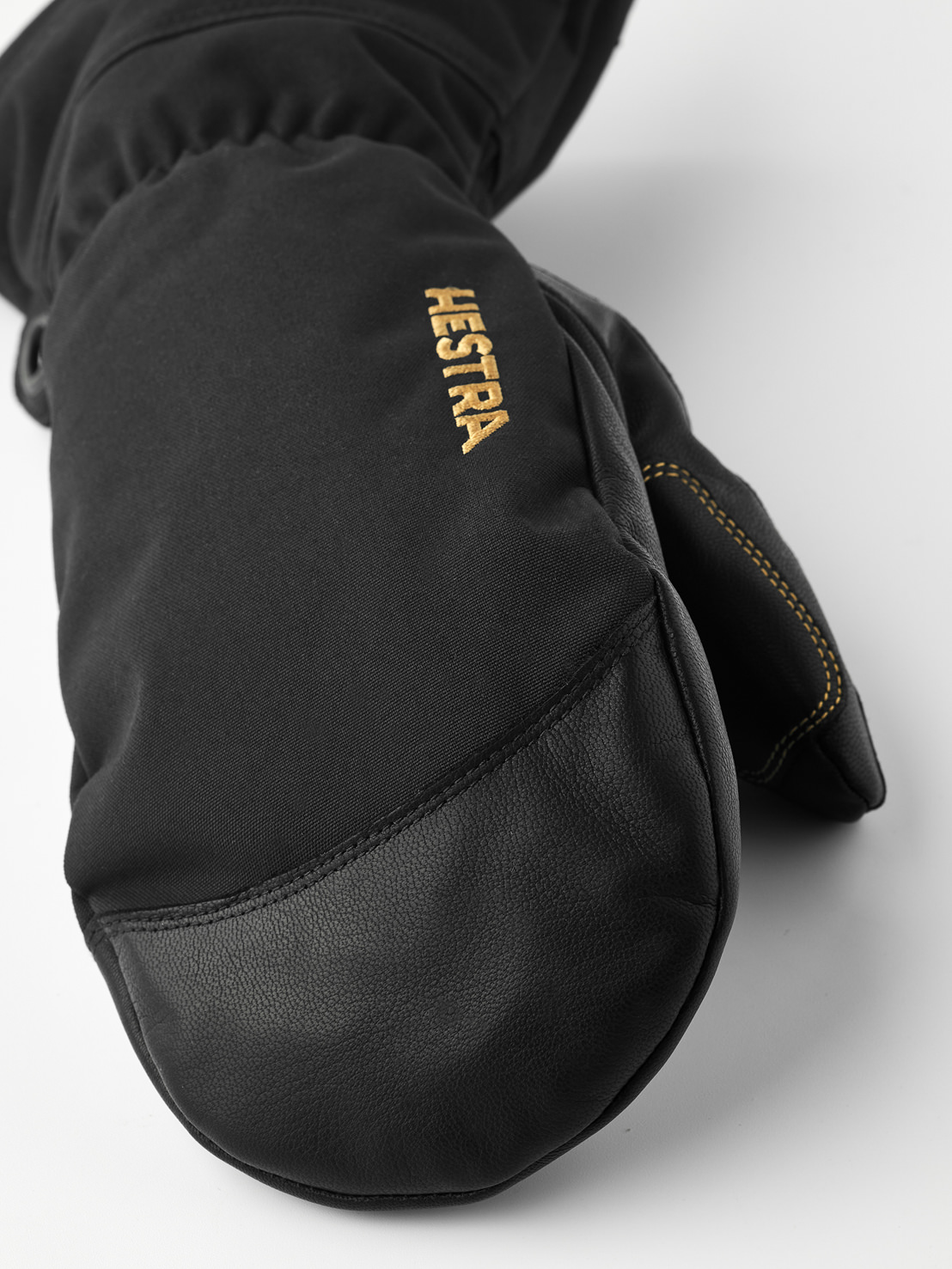 Hestra Fäustling Schwarz Detailaufnahme eines schwarzen Hestra Fäustlings aus Leder und Stoff, mit dem goldfarbenen Hestra-Logo auf dem Handrücken.