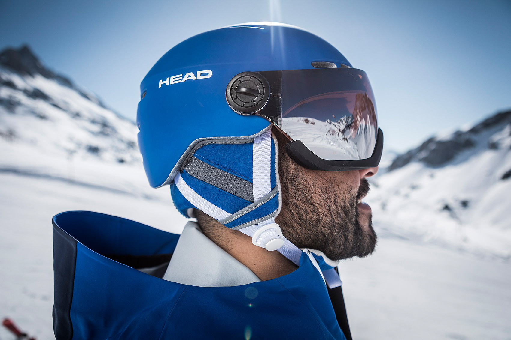Die Vorteile vom Skihelm mit Visier für Brillenträger Profilansicht eines Mannes mit blauem Skihelm und Visier vor schneebedeckter Bergkulisse.