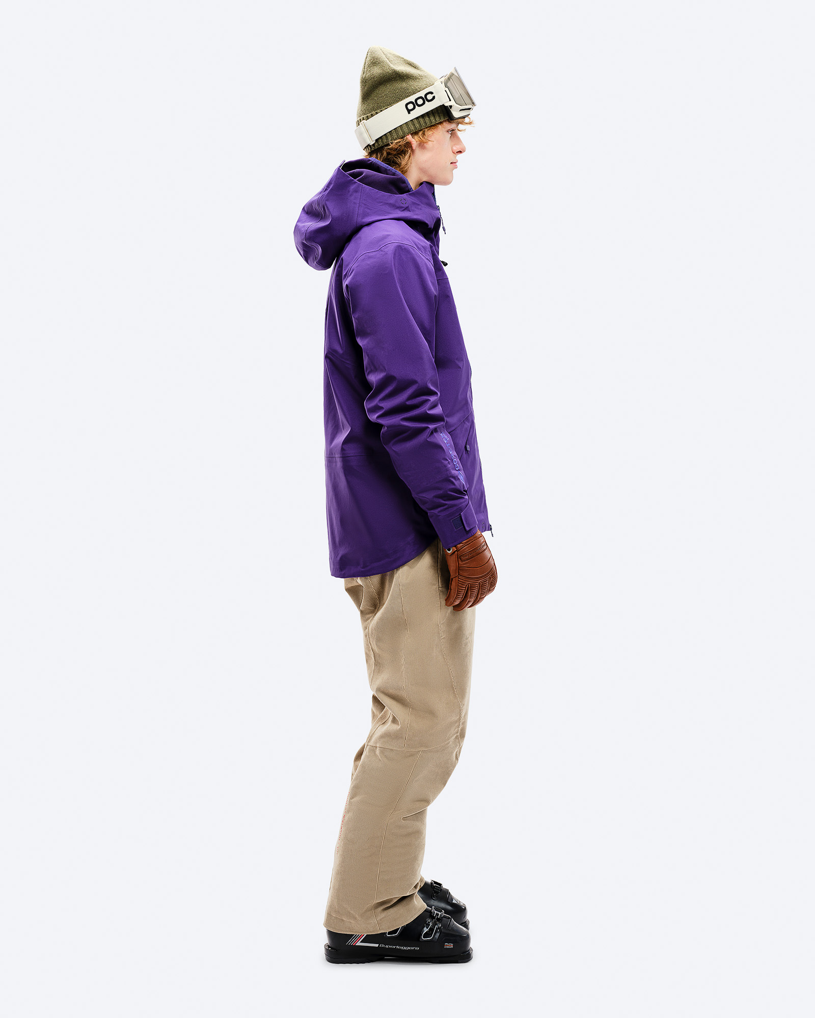 Lila GORE-TEX Jacke Person von der Seite in lila Z-2 GORE-TEX Jacke, beigen Hosen, Skischuhen, Mütze und Brille vor weißem Hintergrund.