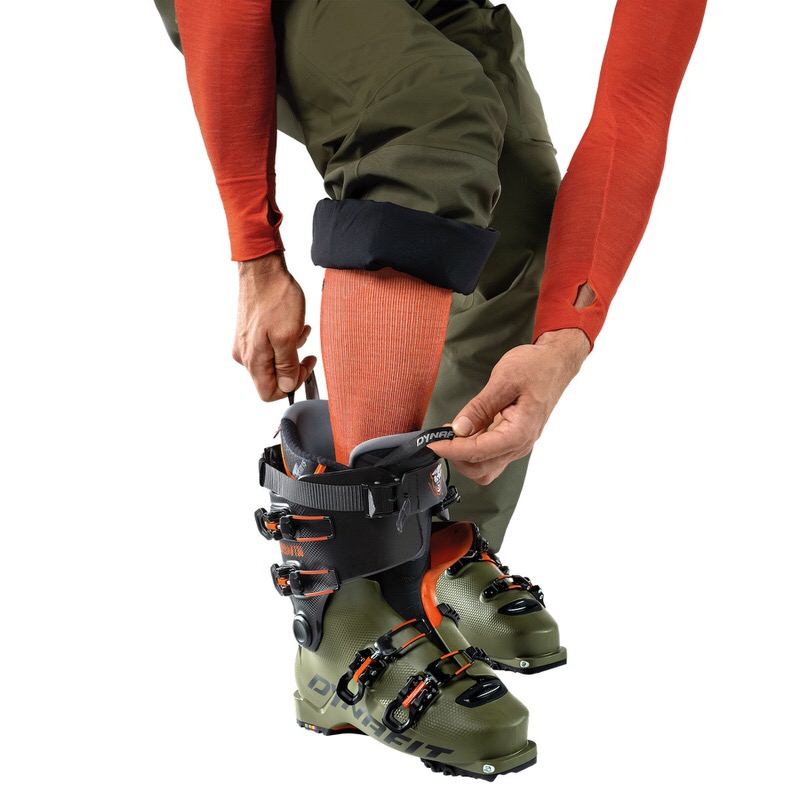 Beheizbare Socken oder Sohlen? Person zieht Dynafit Tigard Alpine Tech Tourenskischuh an. Der Skischuh ist grün und schwarz mit orangefarbenen Akzenten.