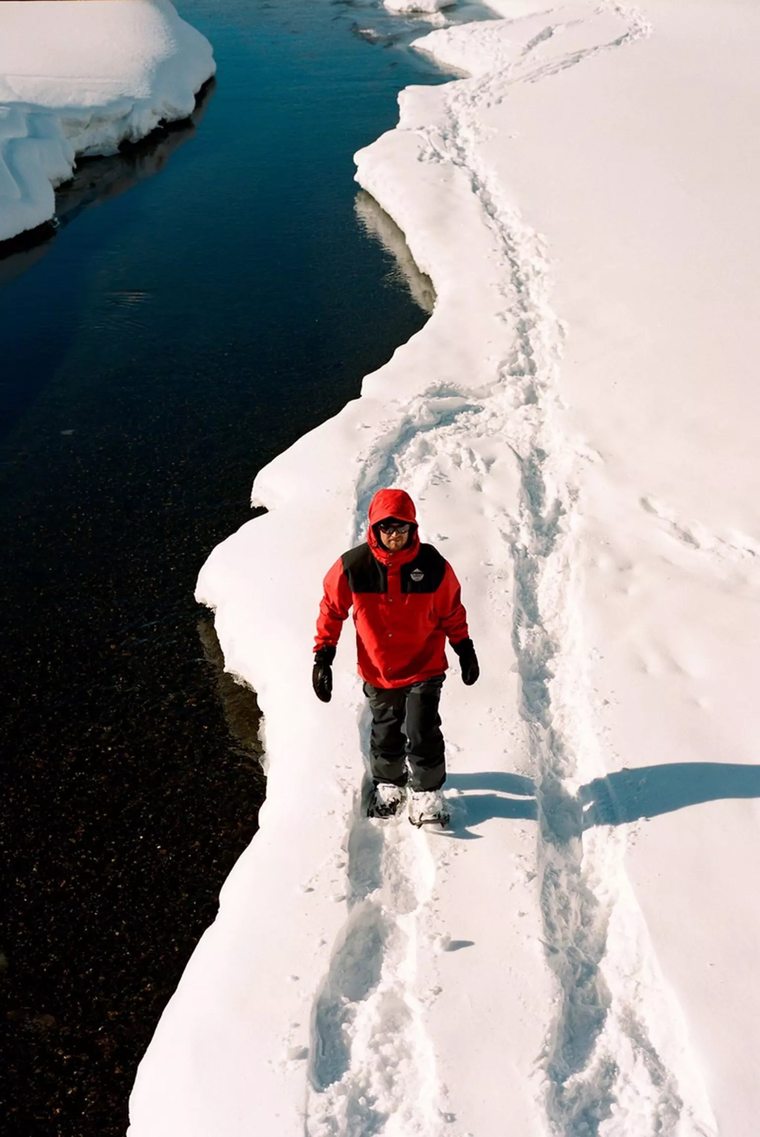 Mann wandert im Schnee Mann mit Tubbs Flex Alp Schneeschuhen wandert auf einem Schneepfad neben einem Fluss. Rote Jacke.