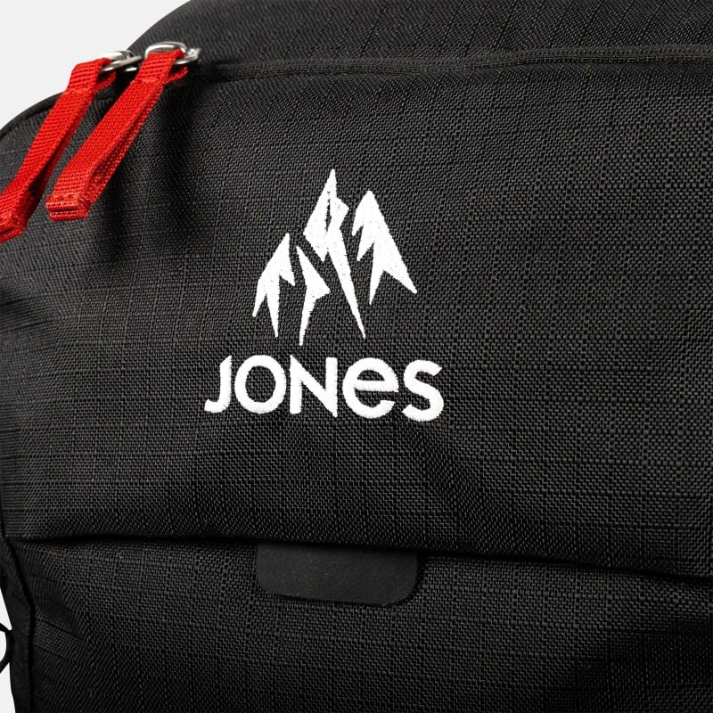 Schwarzer Jones Further Rucksack Detailansicht eines schwarzen Jones Further 25L Rucksacks mit weißem Logo und roten Reißverschlüssen.