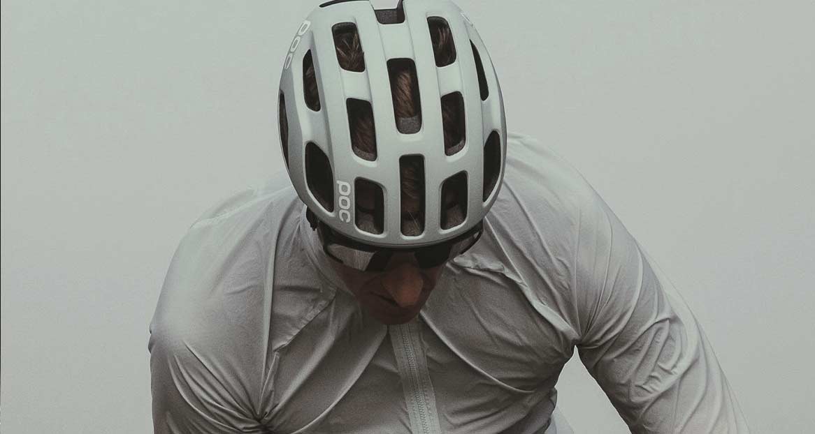 Ein Mann mit Sonnenbrille und weißem Velohelm der Marke POC schaut nach unten. Er trägt eine helle Jacke.
