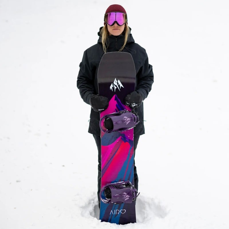 Jones Airheart 2.0 Snowboard Frau mit Skibrille hält Jones Airheart 2.0 Snowboard mit farbenfrohem Design im Schnee.
